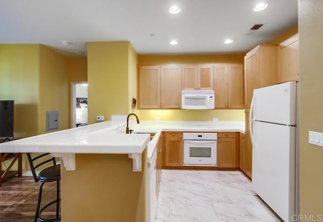 kitchen featuring light brown cabinetry, a kitchen breakfast bar, a sink, white appliances, and a peninsula