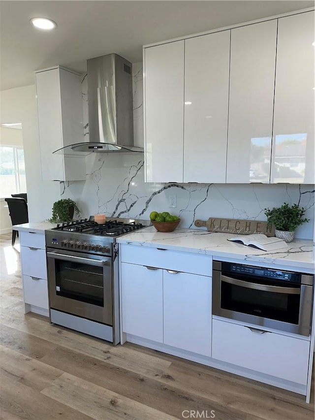 kitchen featuring wall chimney exhaust hood, light wood-type flooring, appliances with stainless steel finishes, light stone counters, and white cabinetry