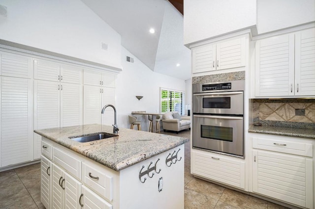 kitchen with high vaulted ceiling, white cabinets, sink, an island with sink, and double oven