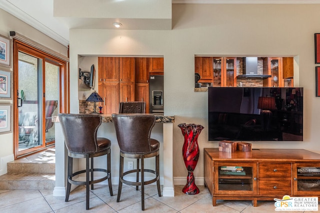 kitchen featuring stainless steel fridge, light tile patterned flooring, wall chimney range hood, and a breakfast bar area