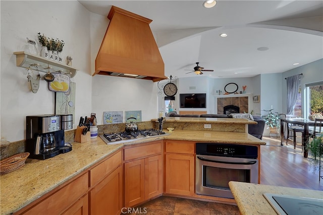 kitchen with appliances with stainless steel finishes, a tile fireplace, ceiling fan, custom range hood, and dark hardwood / wood-style floors