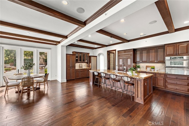 kitchen featuring decorative backsplash, a large island with sink, beamed ceiling, dark hardwood / wood-style floors, and oven