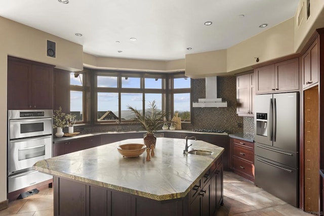 kitchen with sink, a kitchen island with sink, stainless steel appliances, wall chimney range hood, and decorative backsplash