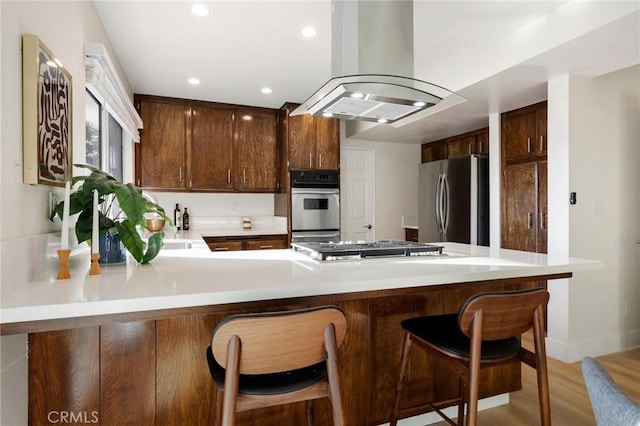 kitchen with island exhaust hood, appliances with stainless steel finishes, light wood-type flooring, kitchen peninsula, and a breakfast bar