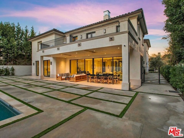 back house at dusk featuring an outdoor living space, a balcony, a fenced in pool, and a patio