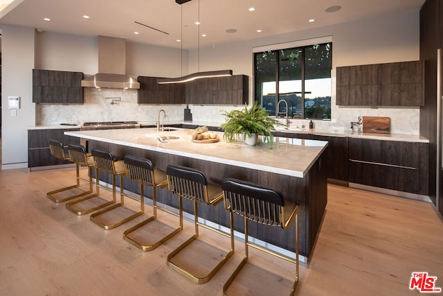 kitchen with light wood-type flooring, backsplash, wall chimney range hood, a center island with sink, and a breakfast bar area