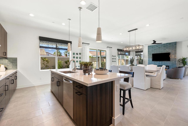 kitchen featuring dishwasher, backsplash, a tiled fireplace, dark brown cabinetry, and a kitchen island with sink