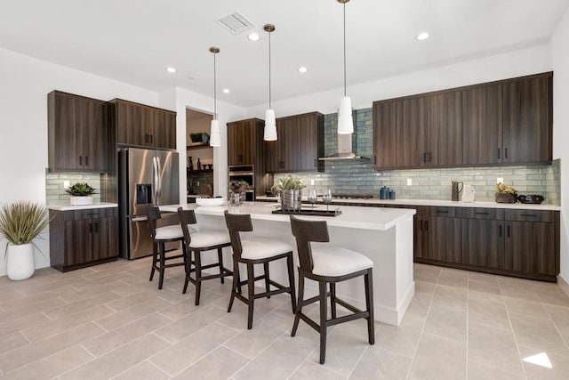 kitchen with wall chimney range hood, dark brown cabinets, hanging light fixtures, an island with sink, and appliances with stainless steel finishes