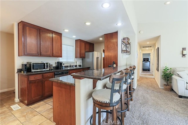 kitchen featuring stainless steel appliances, kitchen peninsula, dark stone countertops, light colored carpet, and a breakfast bar