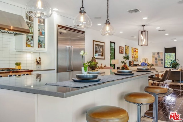 kitchen featuring decorative backsplash, hanging light fixtures, stainless steel appliances, range hood, and white cabinets