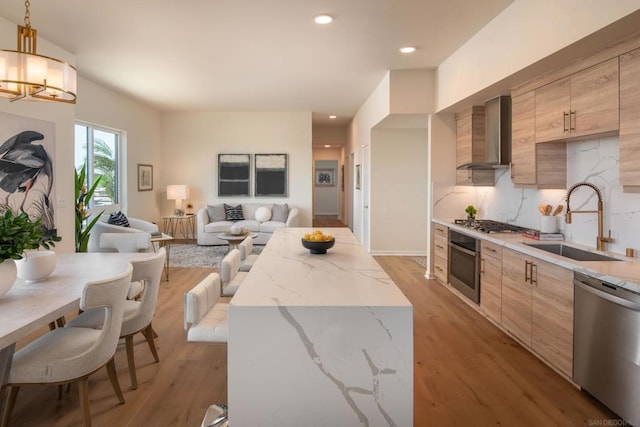 kitchen featuring light wood-type flooring, stainless steel appliances, sink, wall chimney range hood, and decorative light fixtures