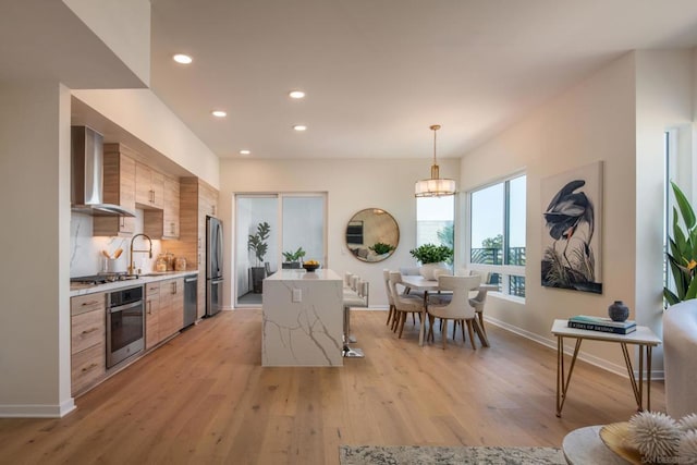 kitchen with wall chimney range hood, decorative backsplash, decorative light fixtures, light hardwood / wood-style floors, and stainless steel appliances