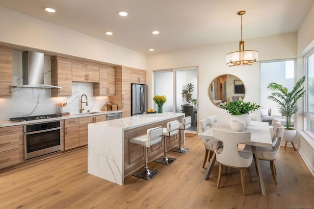 kitchen featuring sink, wall chimney exhaust hood, hanging light fixtures, stainless steel appliances, and light hardwood / wood-style flooring