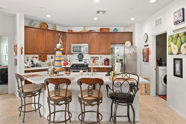 kitchen with washer / dryer, stainless steel appliances, a kitchen island with sink, and light tile patterned flooring