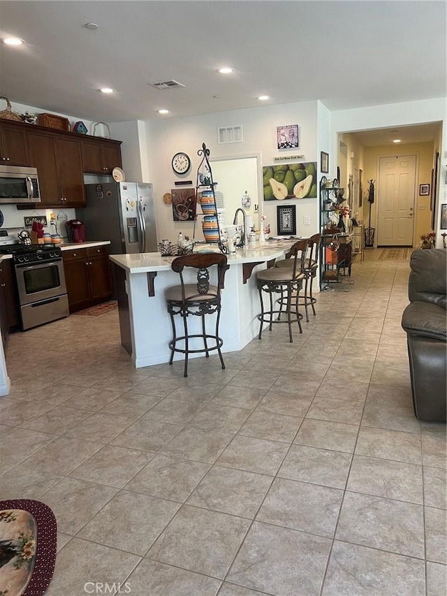 kitchen with dark brown cabinetry, stainless steel appliances, a kitchen breakfast bar, a center island with sink, and light tile patterned flooring