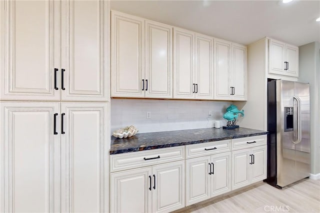 kitchen featuring stainless steel refrigerator with ice dispenser, decorative backsplash, dark stone countertops, light hardwood / wood-style floors, and white cabinetry