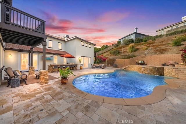 pool at dusk featuring pool water feature, exterior kitchen, an outdoor bar, and a patio