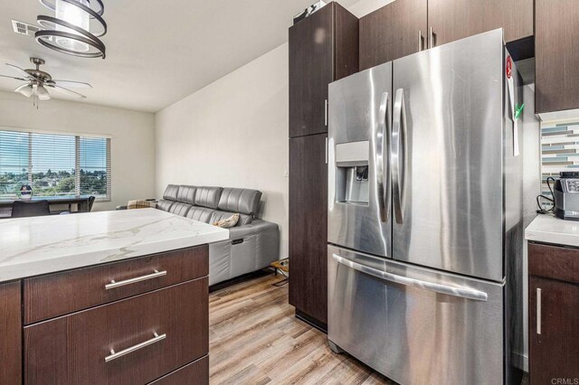 kitchen with stainless steel refrigerator with ice dispenser, dark brown cabinetry, light wood-type flooring, light stone counters, and ceiling fan