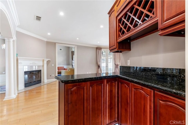 kitchen with dark stone countertops, light wood-type flooring, ornamental molding, ornate columns, and kitchen peninsula