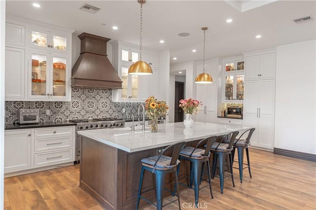 kitchen with white cabinetry, light hardwood / wood-style flooring, premium range hood, an island with sink, and pendant lighting