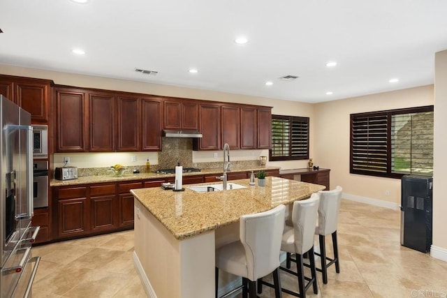 kitchen with an island with sink, a kitchen breakfast bar, light stone countertops, stainless steel appliances, and sink