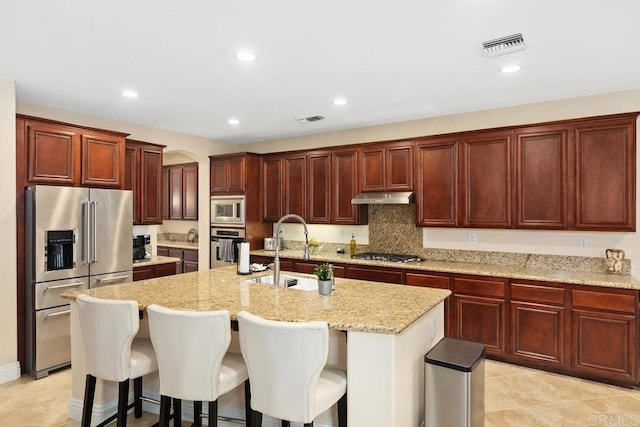 kitchen with a kitchen breakfast bar, an island with sink, stainless steel appliances, sink, and light stone countertops