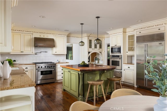 kitchen featuring hanging light fixtures, a center island with sink, custom range hood, built in appliances, and butcher block countertops