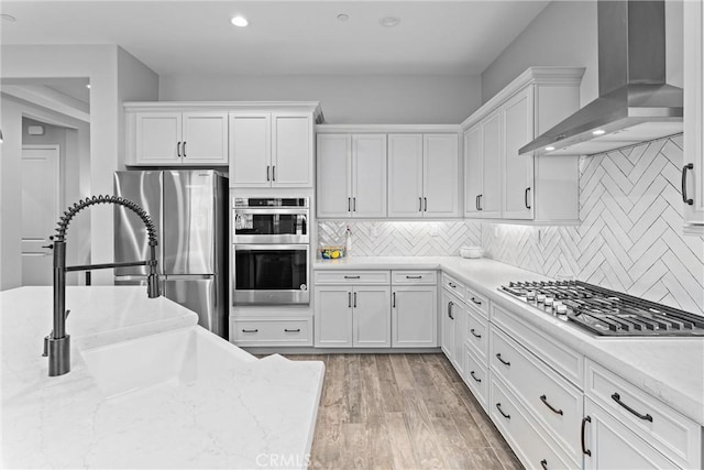 kitchen featuring white cabinetry, wall chimney range hood, light hardwood / wood-style floors, decorative backsplash, and appliances with stainless steel finishes