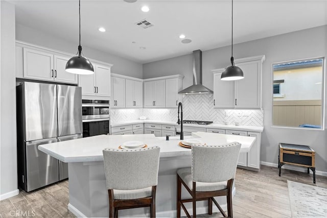 kitchen featuring appliances with stainless steel finishes, a center island with sink, hanging light fixtures, and wall chimney exhaust hood