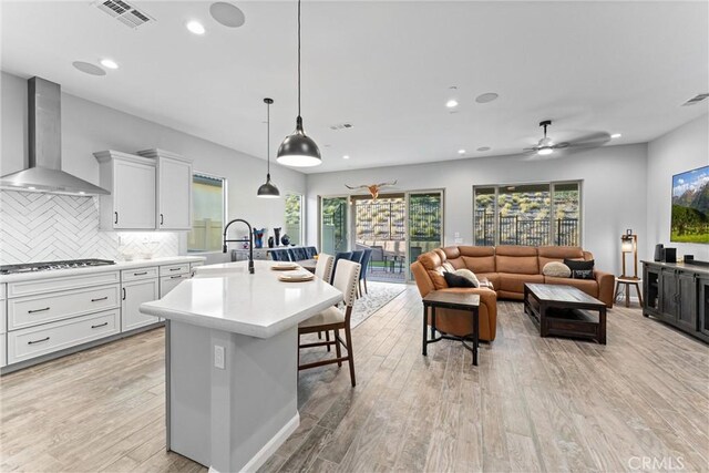 kitchen with a center island with sink, wall chimney range hood, hanging light fixtures, white cabinetry, and stainless steel gas cooktop