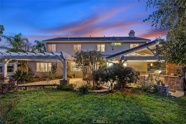 back of property at dusk featuring a patio, stucco siding, a lawn, a pergola, and exterior kitchen