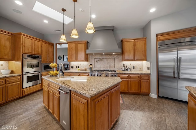 kitchen with an island with sink, wood-type flooring, sink, custom exhaust hood, and appliances with stainless steel finishes