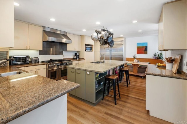 kitchen with an island with sink, wall chimney range hood, high end appliances, a breakfast bar, and sink