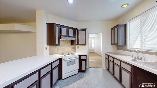 kitchen with white gas range, dark brown cabinetry, tile counters, sink, and backsplash