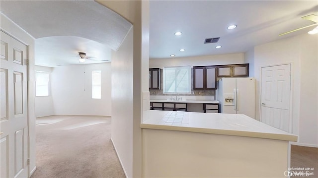 kitchen with ceiling fan, white refrigerator with ice dispenser, kitchen peninsula, light colored carpet, and dark brown cabinets