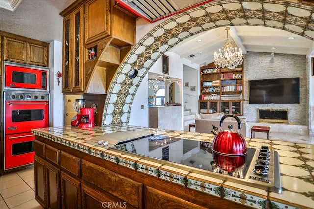 kitchen featuring a fireplace, multiple ovens, lofted ceiling with beams, light tile patterned flooring, and an inviting chandelier