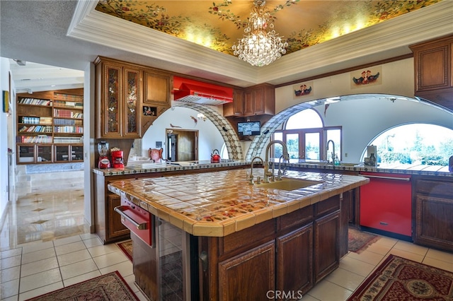 kitchen with sink, a raised ceiling, a kitchen island with sink, tile countertops, and crown molding