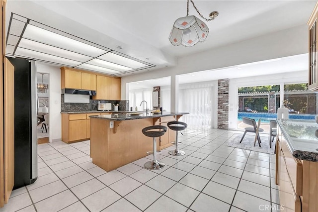 kitchen featuring light brown cabinets, backsplash, a kitchen island with sink, light tile patterned floors, and stainless steel refrigerator