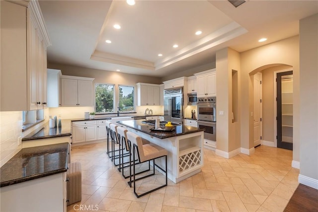 kitchen with a raised ceiling, built in appliances, white cabinetry, and a kitchen island