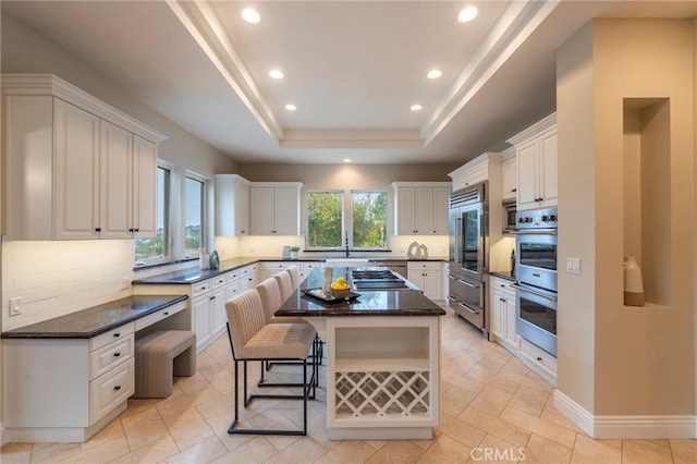 kitchen featuring decorative backsplash, dark stone counters, a tray ceiling, white cabinets, and a center island