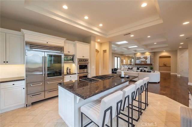 kitchen with light wood-type flooring, dark stone counters, a tray ceiling, built in appliances, and white cabinetry