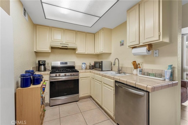 kitchen with tile countertops, cream cabinets, sink, light tile patterned floors, and stainless steel appliances