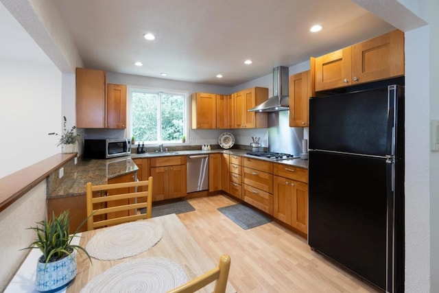 kitchen with sink, wall chimney range hood, light hardwood / wood-style flooring, dark stone countertops, and appliances with stainless steel finishes