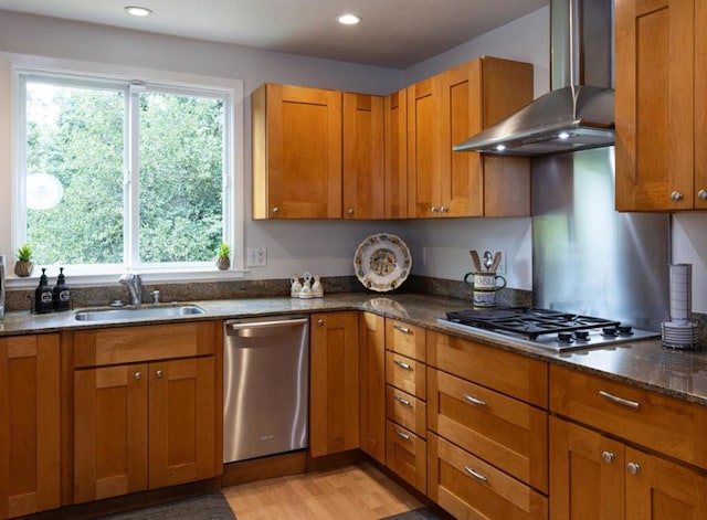 kitchen with appliances with stainless steel finishes, wall chimney exhaust hood, sink, light hardwood / wood-style flooring, and dark stone countertops