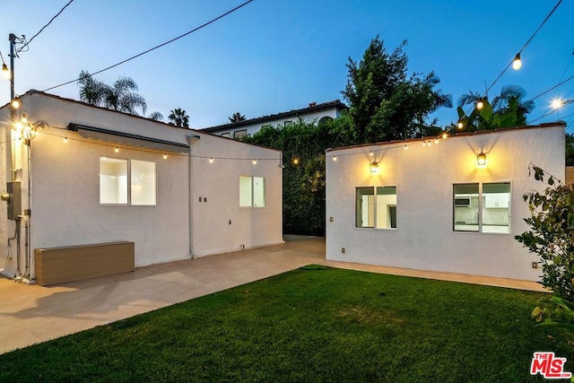 back house at dusk featuring a lawn and a patio