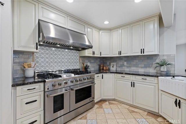 kitchen featuring decorative backsplash, dark stone counters, wall chimney exhaust hood, and range with two ovens