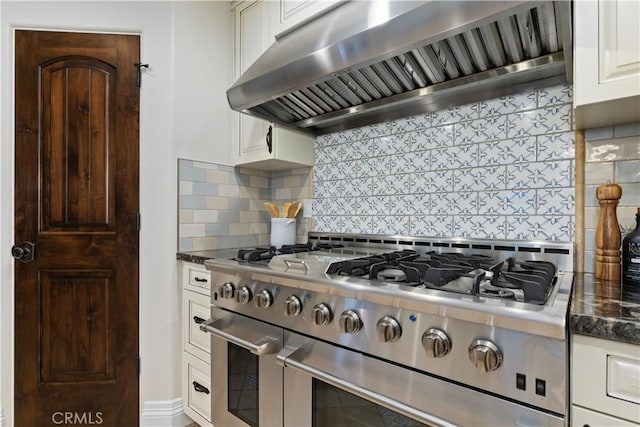 kitchen with wall chimney range hood, dark stone countertops, white cabinets, decorative backsplash, and range with two ovens