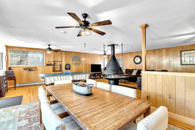 dining room featuring a baseboard radiator, light wood-type flooring, a wood stove, ceiling fan, and wooden walls