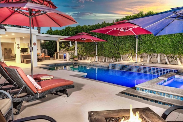 pool at dusk featuring an in ground hot tub, a patio area, an outdoor kitchen, exterior fireplace, and a fire pit