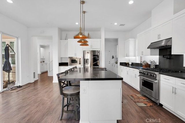 kitchen with stainless steel appliances, a kitchen island with sink, dark wood-type flooring, decorative light fixtures, and white cabinets